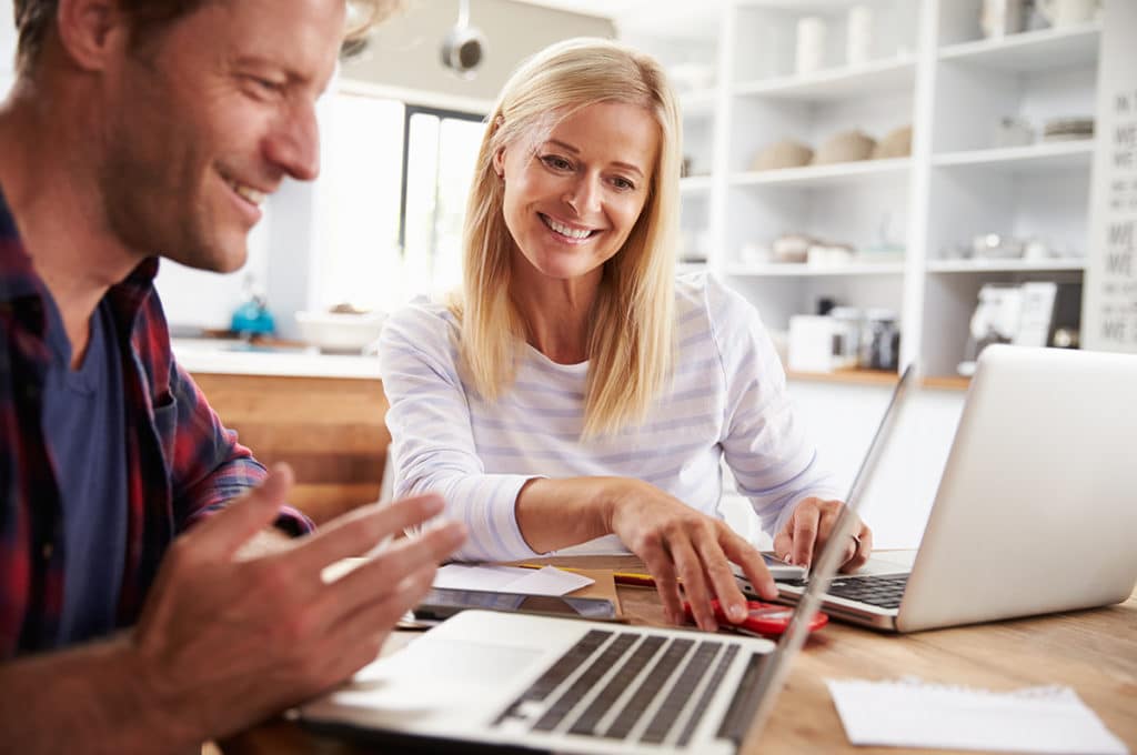 A couple smiling and looking at their finances on their laptops