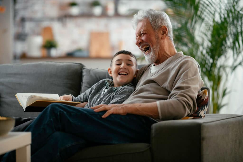 Happy Grandfather and Grandson sitting on a couch, reading a book, and laughing.